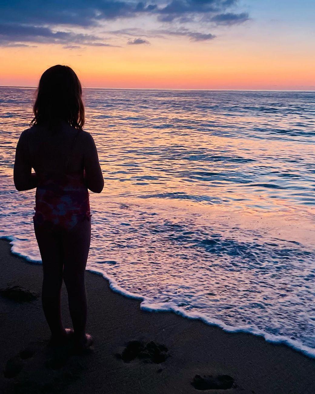 A child standing at the water&rsquo;s edge, watching the sunset over the sea